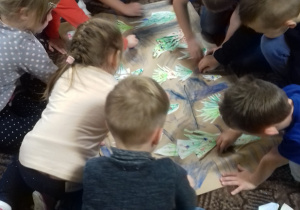 grupa dzieci kucają na dywanie i wykonują wspólną pracę na szarym papierze